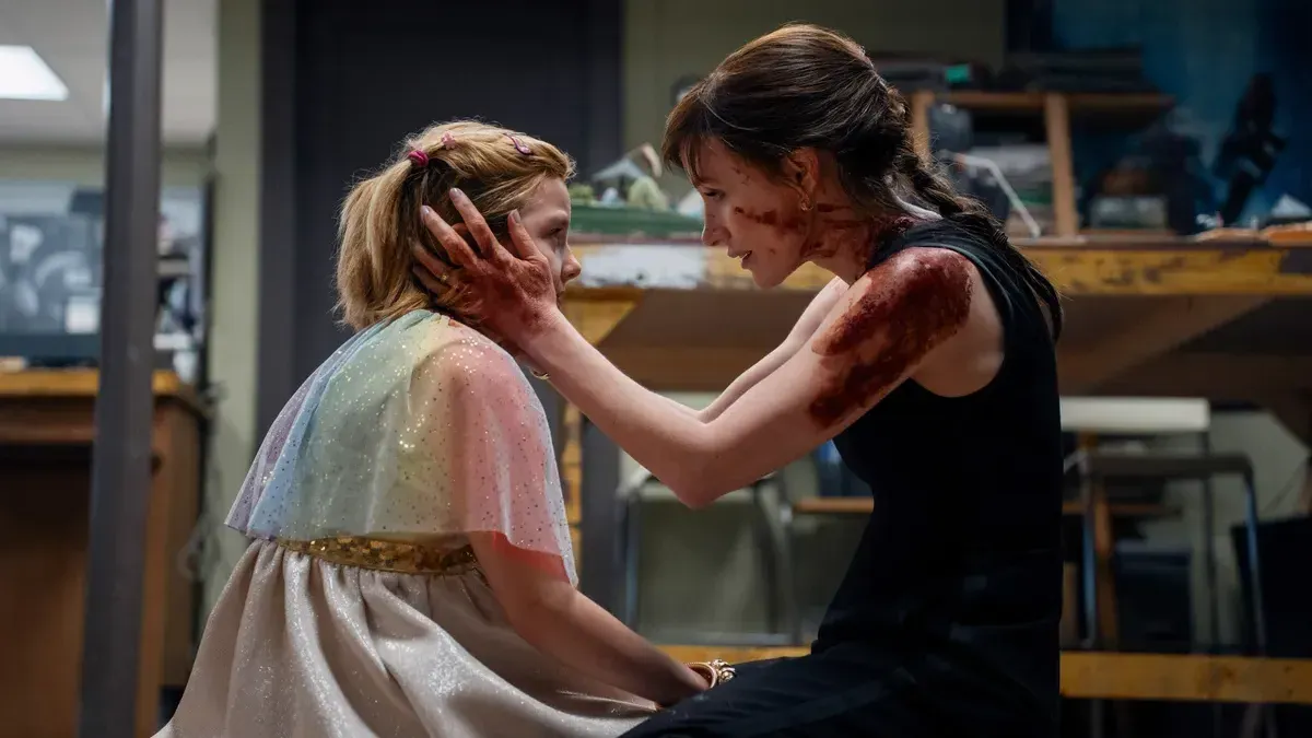 A woman with blood on her face and arms kneels and holds a young girl’s face in her hands in a cluttered basement, in a still from Other Mommy