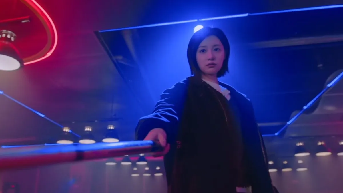 A woman in a dark jacket points a pool cue towards the camera under blue and pink neon light, in a still from Doctor X: Mafia in White