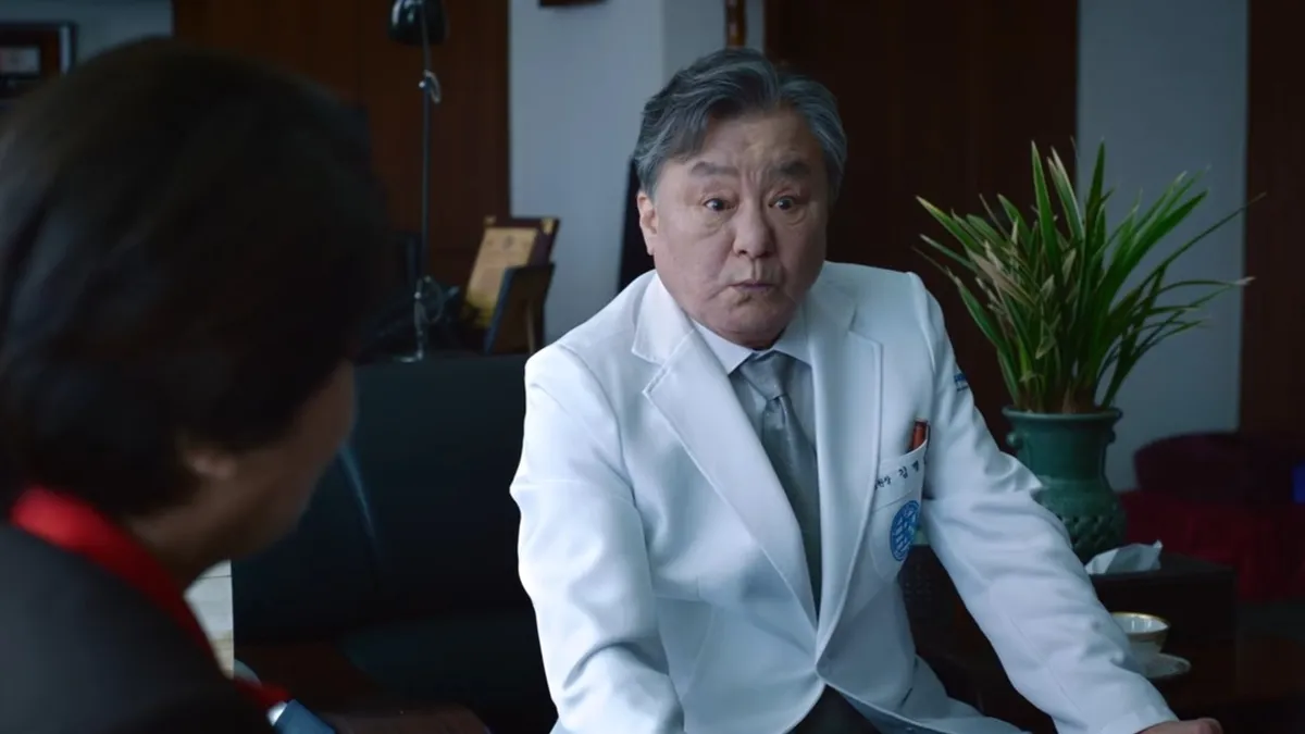 An older doctor in a white coat looks startled at a visitor seated across from him in his office, in a still from Doctor X: Mafia in White