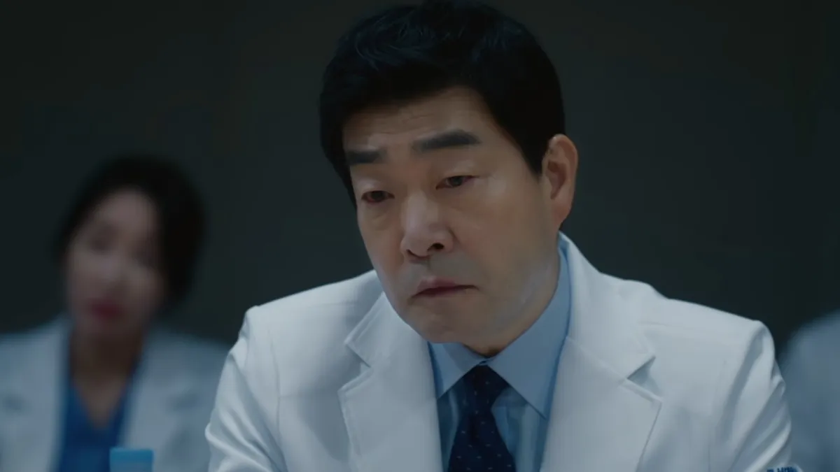 A doctor in a white coat and patterned tie looks down during a meeting, with colleagues out of focus behind him, in a still from Doctor X: Mafia in White