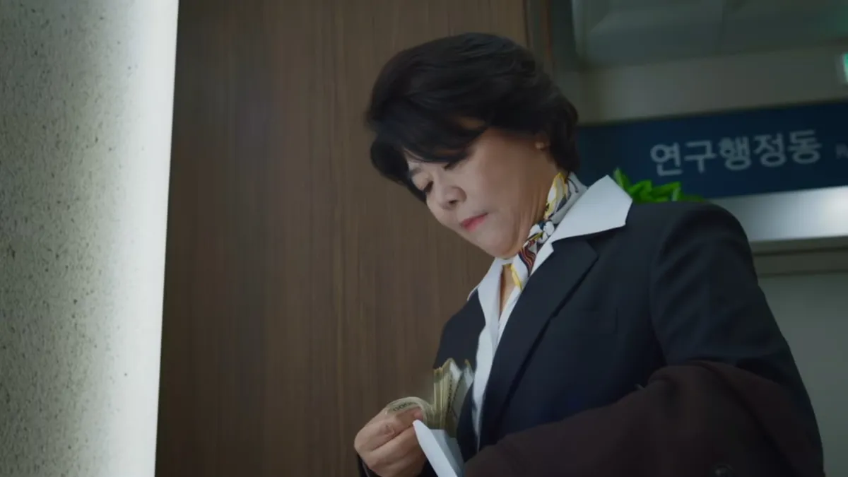 A woman in a dark suit and patterned scarf counts a bundle of cash from an envelope in a hospital corridor, in a still from Doctor X: Mafia in White