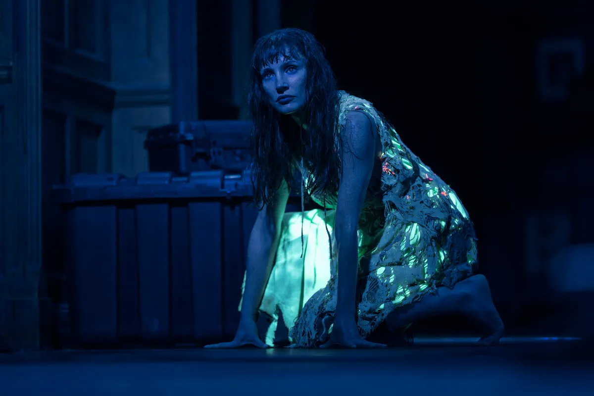 A long-haired figure in a pale dress crouches on all fours in a dark blue room, light glowing beneath it, in a still from Other Mommy