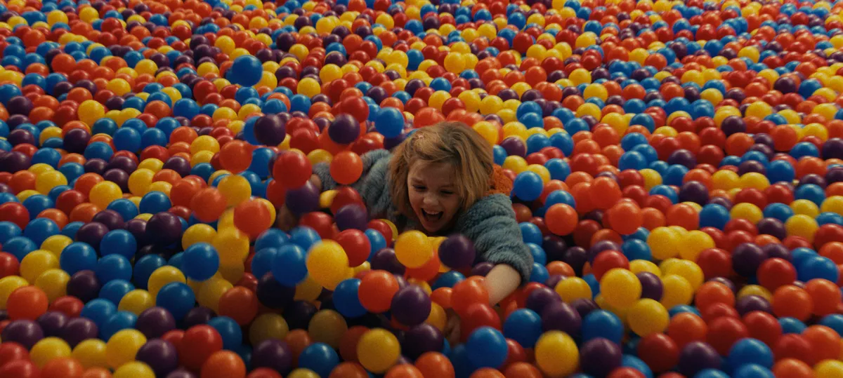 A girl lies on her front in a huge pit of red, yellow and blue plastic balls, reaching forward, in a still from Other Mommy