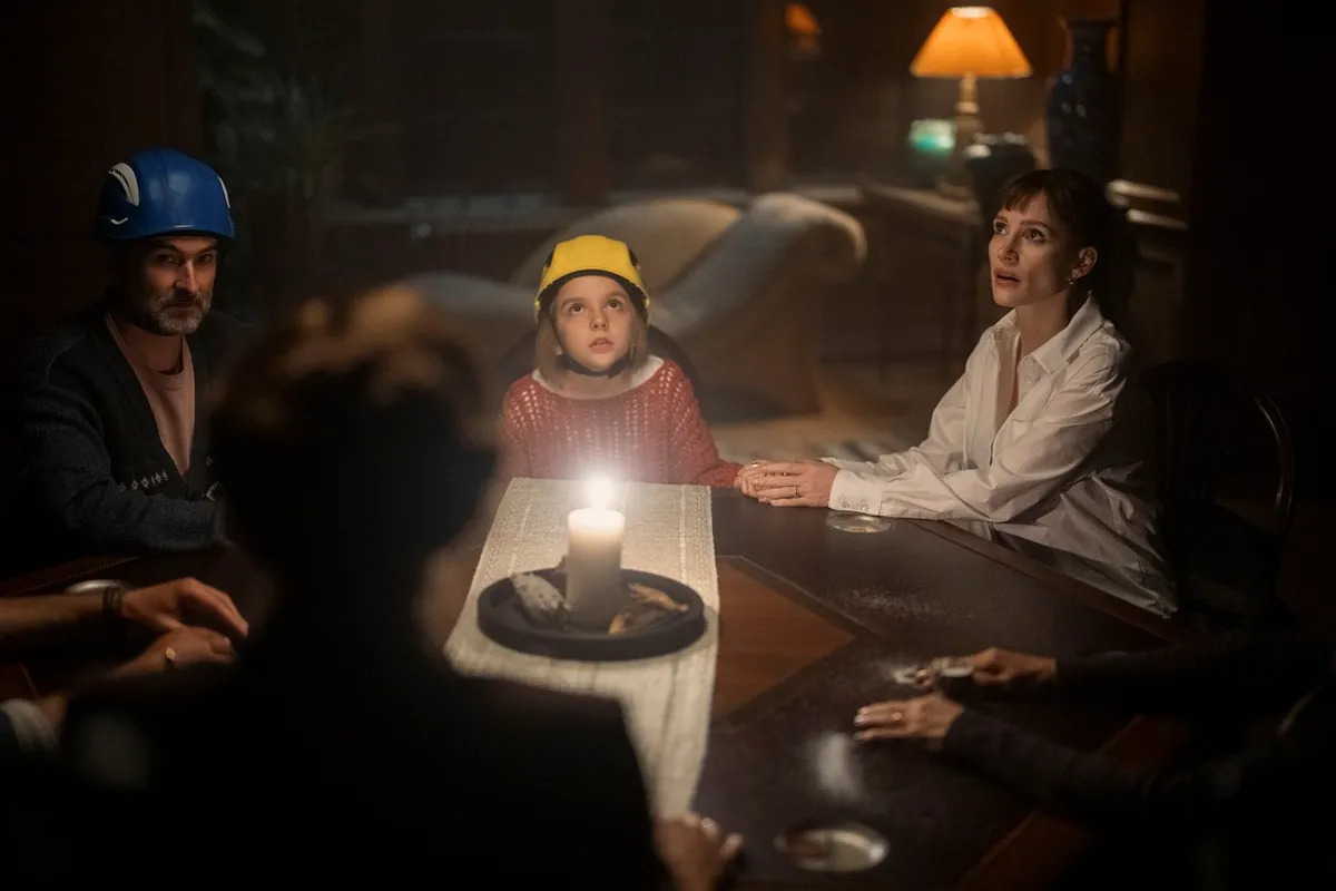 A child in a yellow hat sits at a candlelit table between a man in a blue helmet and a woman in a white shirt, in a still from Other Mommy