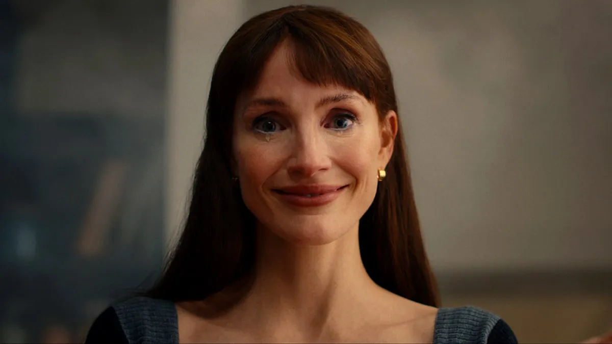 A woman with long red hair and a fringe gives a wide, fixed smile in close-up, in a still from Other Mommy